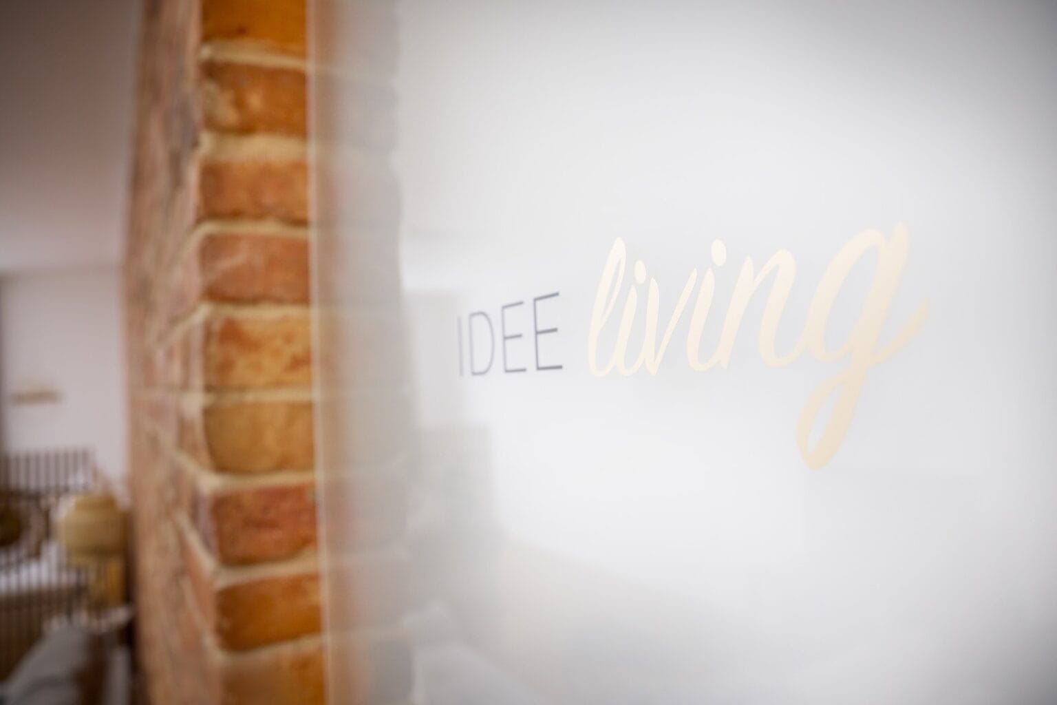 IDEE Living - Eine Nahaufnahme einer Milchglastür mit der Aufschrift „IDEE living“. Durch das Glas sind eine verschwommene Ziegelwand und einige Einrichtungsgegenstände zu sehen.