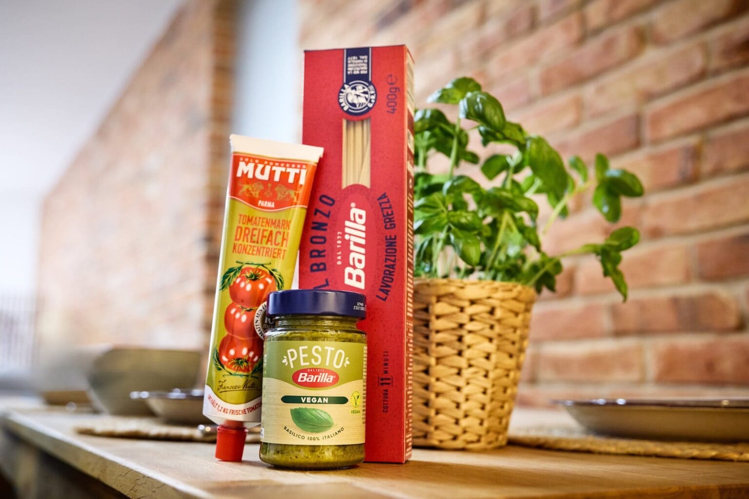 IDEE Living - Eine Tube Tomatenmark, eine Packung Spaghetti und ein Glas Pesto stehen auf einer Holzfläche, im Hintergrund ist ein Topf mit Basilikum zu sehen.