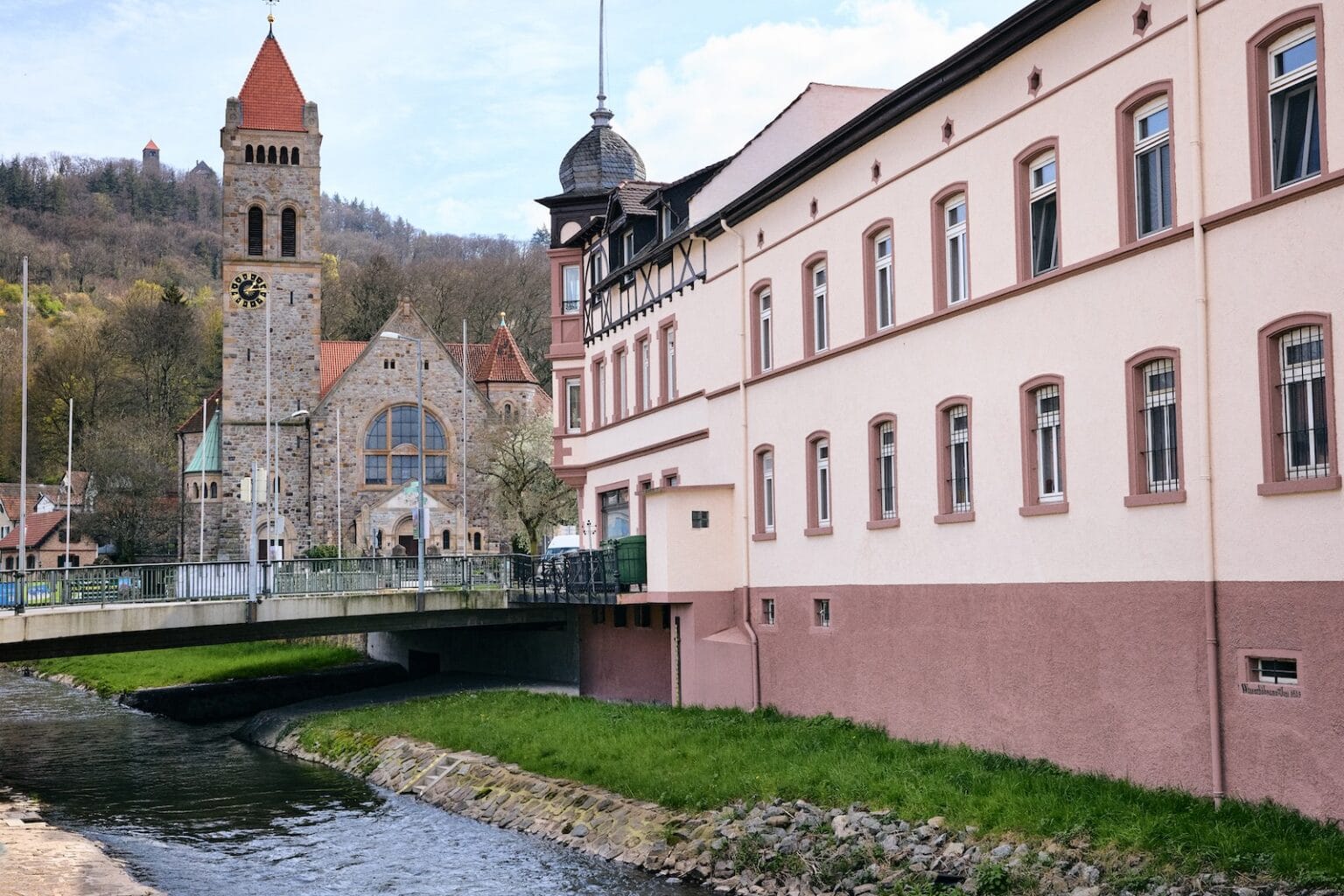 IDEE Living - Ein Bach fließt unter einer Brücke neben einem rosafarbenen Gebäude hindurch. Im Hintergrund ist eine Steinkirche mit einem Glockenturm und einem roten Dach vor einem Hügel mit Bäumen zu sehen.