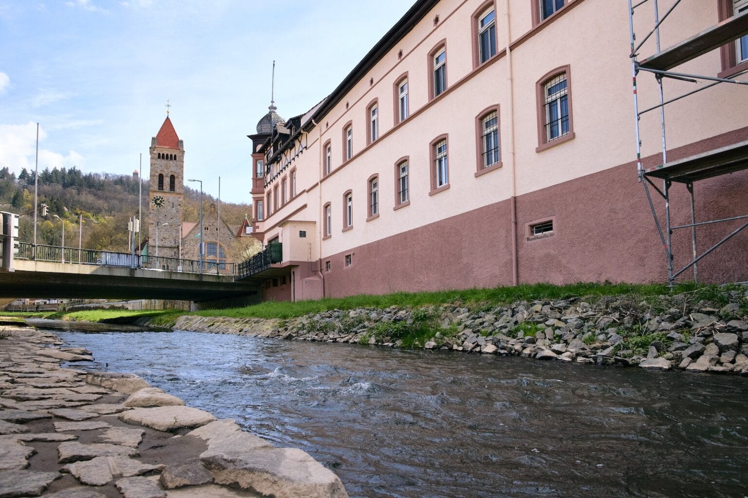 IDEE Living - Ein Fluss fließt neben einem alten Gebäude mit einem Uhrenturm im Hintergrund. Über den Fluss führt eine Brücke, und auf der rechten Seite des Gebäudes ist ein Gerüst zu sehen.