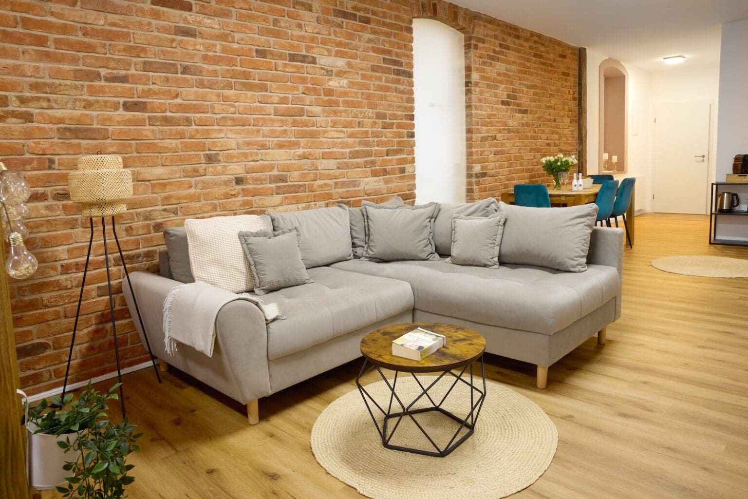 IDEE Living - Ein modernes Wohnzimmer mit einem grauen Sofa, einem runden Couchtisch, Backsteinwänden und einem Essbereich im Hintergrund. Der Raum verfügt über Holzböden und minimalistisches Dekor.