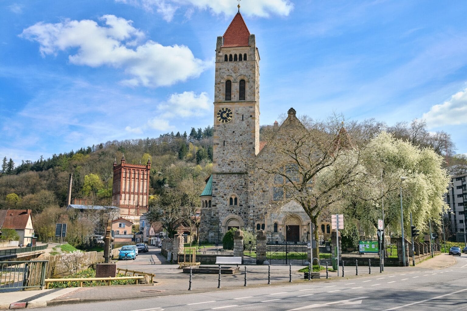 IDEE Living - Eine Steinkirche mit einem Glockenturm steht an einer von Bäumen gesäumten Straße, neben einem teilweise sichtbaren Gasometer und den umliegenden Gebäuden. Im Hintergrund ist ein bewaldeter Hügel zu sehen.