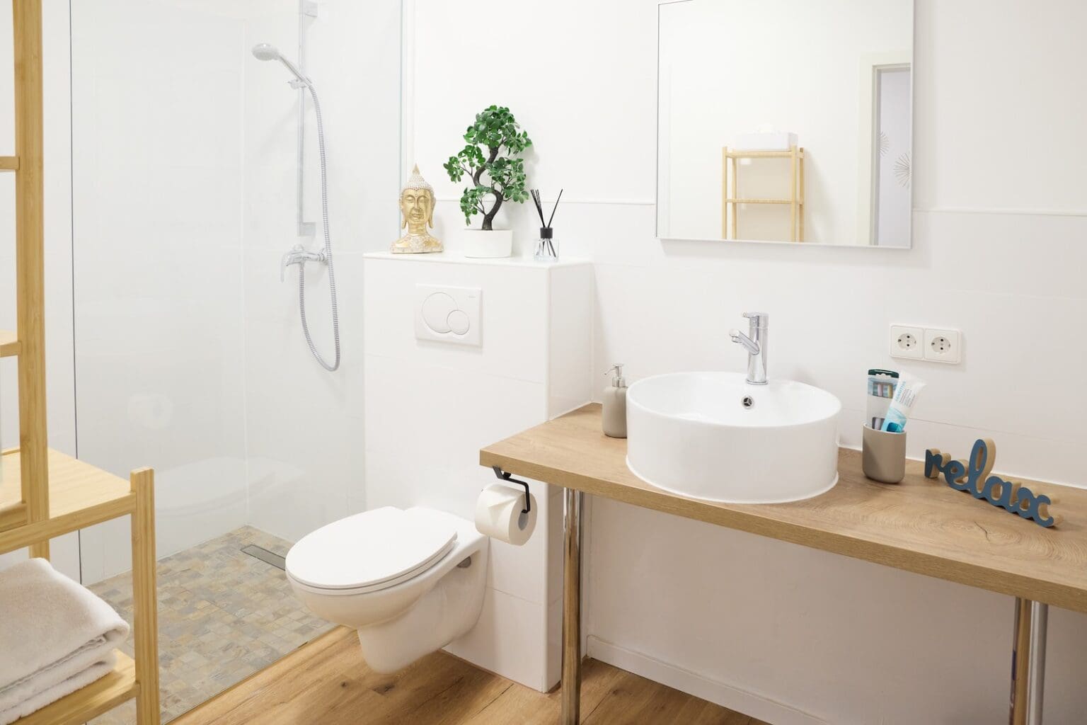 IDEE Living - Modernes Badezimmer mit ebenerdiger Dusche, weißer Toilette und einer hölzernen Arbeitsplatte mit rundem Waschbecken. Zu den Accessoires gehören ein Spiegel, Toilettenartikel, eine kleine Topfpflanze und ein „Relax“-Schild.