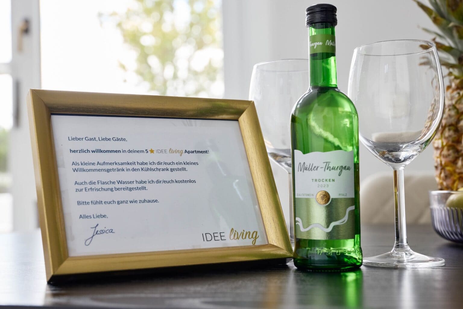 IDEE Living - Eine gerahmte Willkommenskarte, eine Flasche Müller-Thurgau-Wein und zwei leere Weingläser sind neben einer Obstschale auf einem Tisch arrangiert.