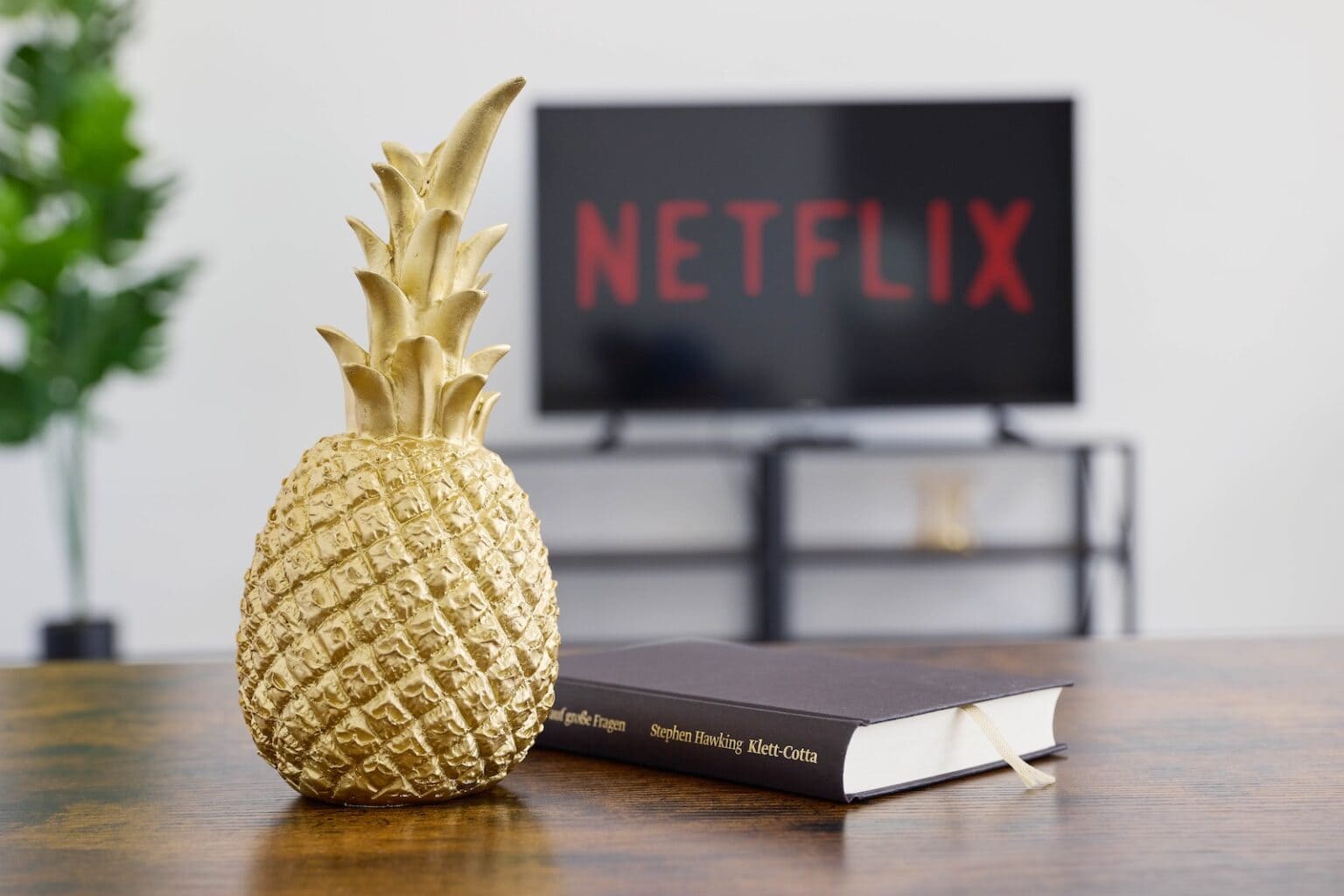 IDEE Living - Eine dekorative goldene Ananas, ein Buch und ein Fernseher, auf dem Netflix läuft, stehen auf einem Holztisch in einer Wohnzimmereinrichtung.