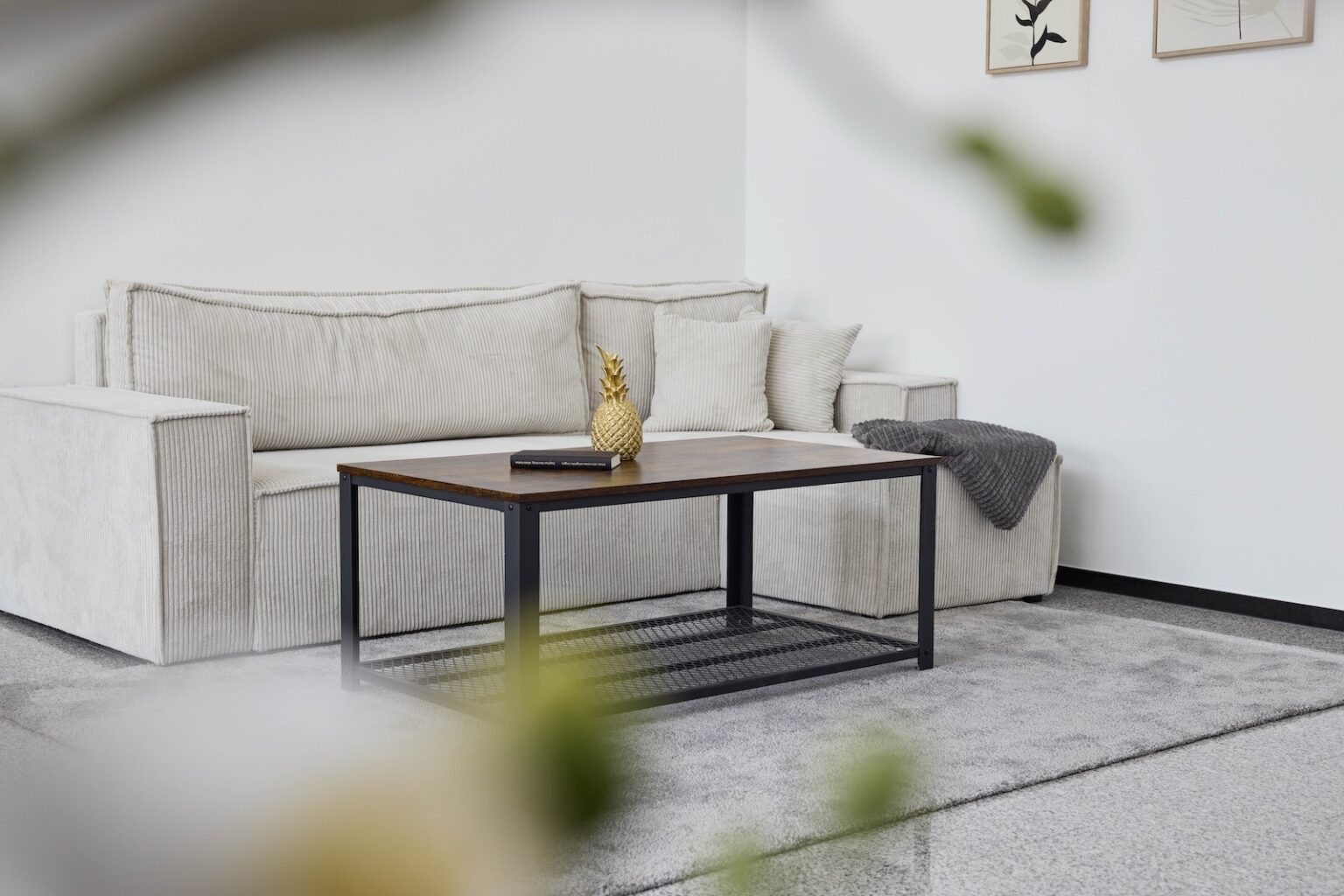 IDEE Living - Ein minimalistisches Wohnzimmer mit einem weißen Sofa, einem Couchtisch aus Holz und einer kleinen goldenen Ananas-Dekoration auf dem Tisch. Über der Armlehne des Sofas hängt eine graue Decke und gerahmte Kunst schmückt die Wand.