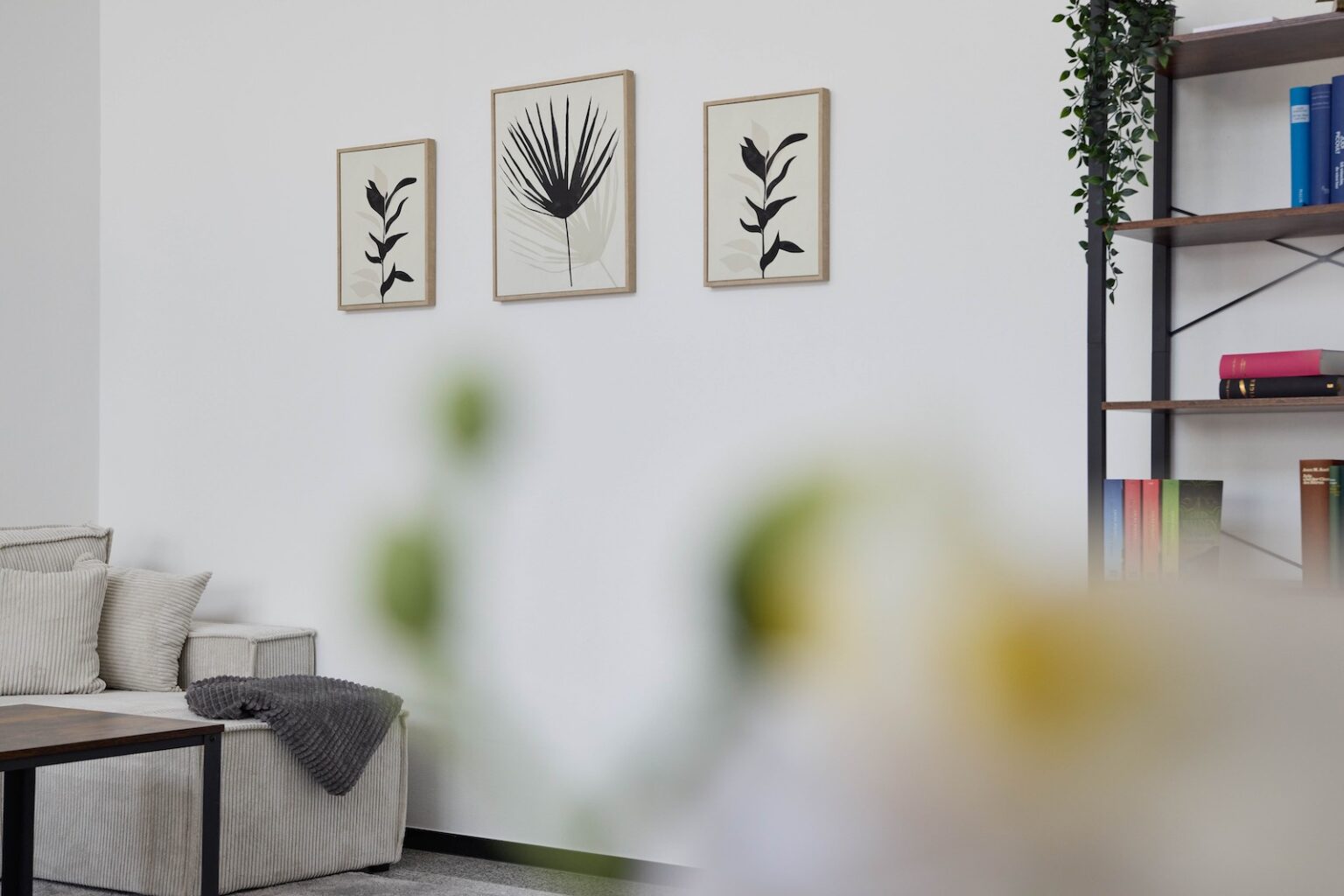 IDEE Living - Ein minimalistisches Wohnzimmer verfügt über ein graues Sofa mit Kissen, einen hölzernen Couchtisch, gerahmte botanische Drucke an der Wand und ein Bücherregal mit Büchern und Pflanzen.