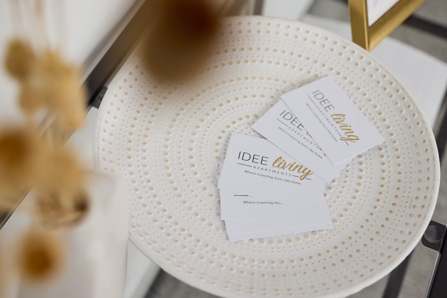 IDEE Living - Auf einem weißen Dekoteller liegen mehrere Visitenkarten von „IDEE Living Apartments“ mit minimalistischem Text in Schwarz und Gold. Die Karten sind ordentlich in einem kleinen Stapel angeordnet.