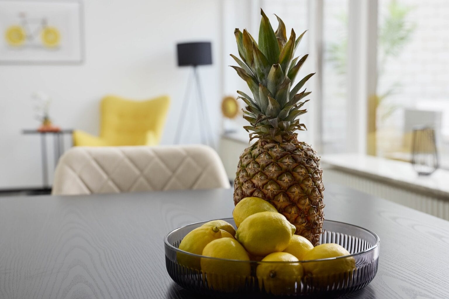 IDEE Living - Eine Ananas und mehrere Zitronen in einer Schale stehen auf einem dunklen Tisch in einem modernen, gut beleuchteten Raum mit einem gelben Stuhl und anderen Möbeln im Hintergrund.