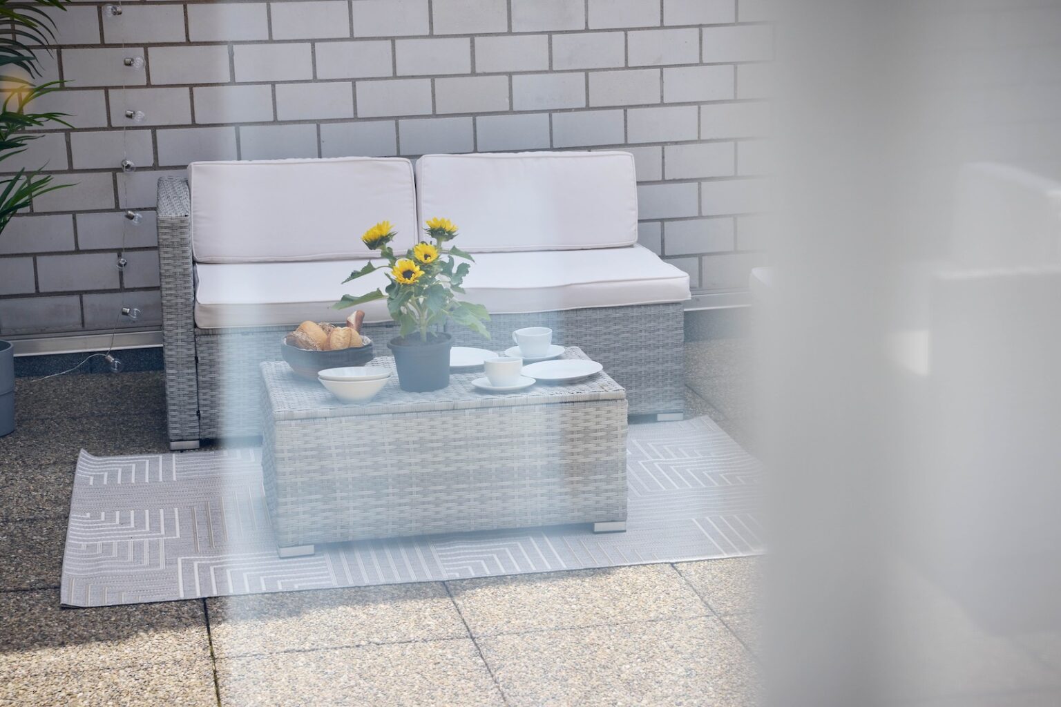 IDEE Living - Eine Terrasse mit einem Korbsofa, einem Couchtisch und Sonnenblumen in Töpfen. Auf dem Tisch stehen Schüsseln, Tassen und Croissants, alles auf einem gemusterten Teppich.