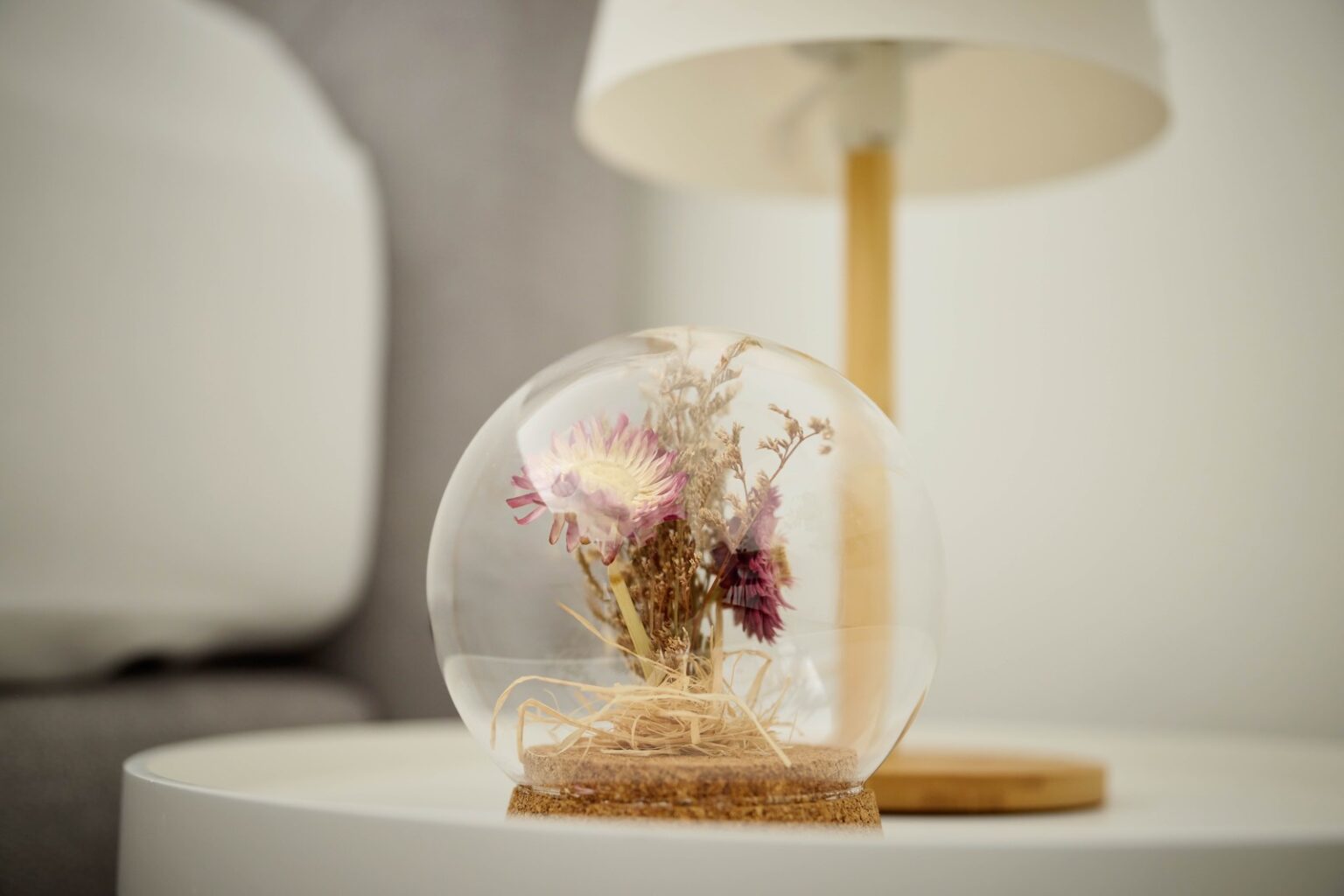 IDEE Living - Eine dekorative Glaskuppel mit getrockneten Blumen auf einem runden weißen Tisch mit einer Lampe im Hintergrund.