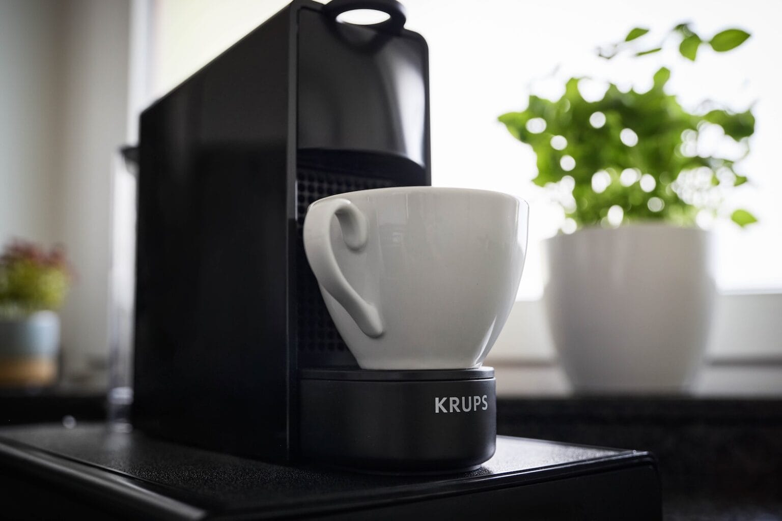 IDEE Living - Eine schwarze Krups-Kaffeemaschine mit einer weißen Tasse steht auf einer Küchentheke, im Hintergrund ist eine Topfpflanze zu sehen.