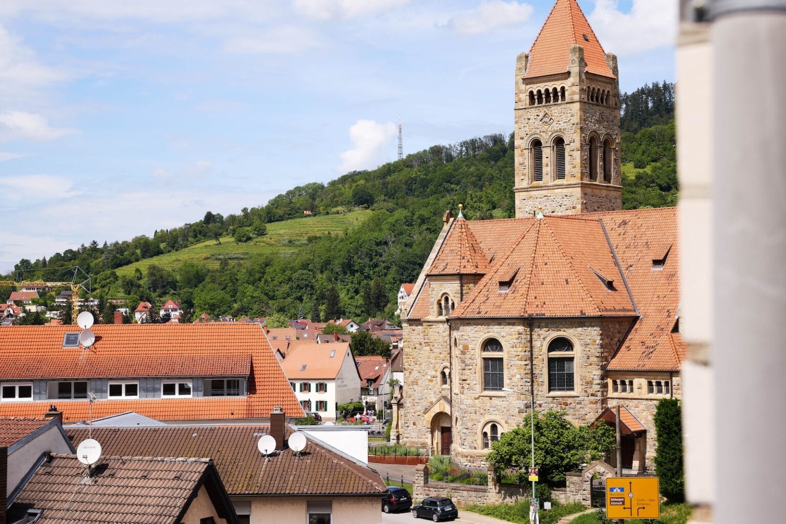 IDEE Living - Eine Kirche mit einem hohen, spitzen Dachturm steht zwischen Wohnhäusern mit orangefarbenen Dächern, vor der Kulisse grüner Hügel und eines teilweise bewölkten Himmels.