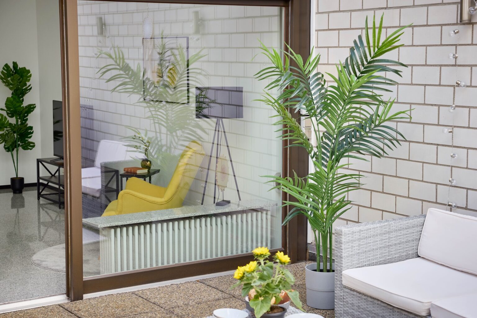 IDEE Living - Eine moderne Terrasse mit einem Korbsofa, Topfpflanzen und einer Glasschiebetür spiegelt einen Wohnraum im Innenbereich mit einem gelben Stuhl, einer Lampe und viel Grün wider.