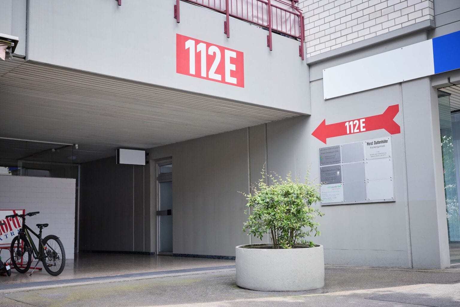 IDEE Living - Eine graue Gebäudefassade mit einem großen roten Schild „112E“. An einer Wand mit Richtungs- und Informationsschildern zeigt ein Pfeil nach rechts auf 112E. In der Nähe sind auch ein Fahrrad und eine Topfpflanze zu sehen.