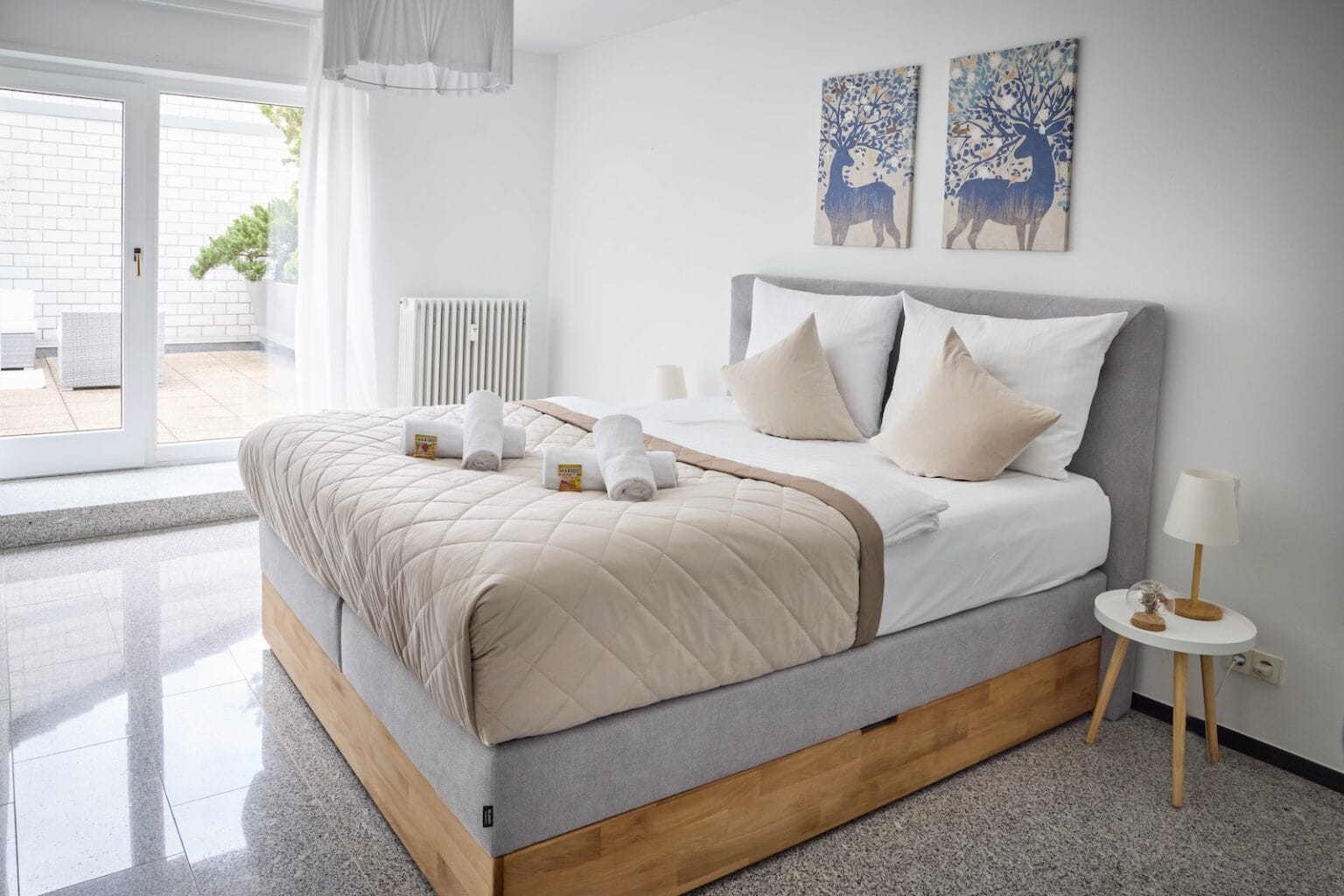 IDEE Living - Ein modernes Schlafzimmer mit einem ordentlich gemachten Bett mit beigen Decken und Kissen, gerollten Handtüchern und zwei kleinen Toilettenartikeln. Das Zimmer verfügt über Wandkunst, einen Beistelltisch mit einer Lampe und eine Glasschiebetür.