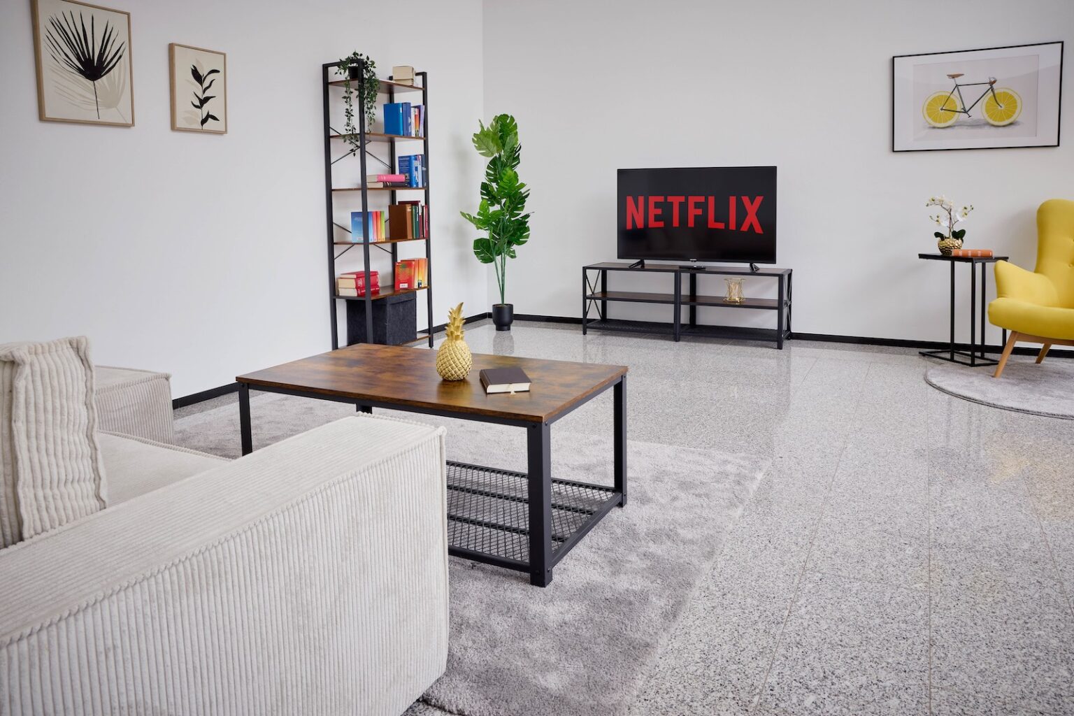 IDEE Living - Ein modernes Wohnzimmer mit einem beigen Sofa, einem hölzernen Couchtisch, einem Fernseher mit „Netflix“, einem Bücherregal und Wandkunst. Ein gelber Stuhl und eine Pflanze sind ebenfalls vorhanden.