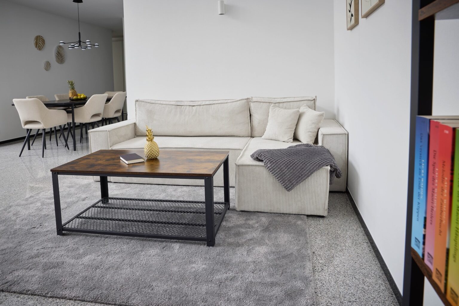 IDEE Living - Ein modernes Wohnzimmer mit einem hellgrauen Sofa, einem hölzernen Couchtisch und einem grauen Teppich. Im Hintergrund ist ein Essbereich mit beigen Stühlen zu sehen. Über dem Sofa liegt eine graue Decke.