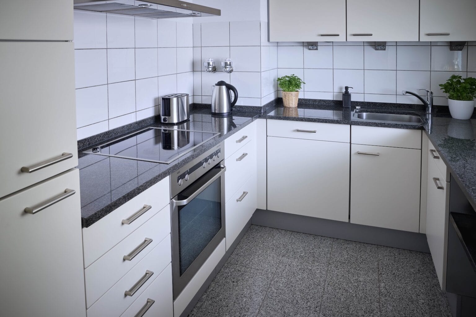 IDEE Living - Eine moderne Küche mit weißen Schränken, Geräten aus Edelstahl einschließlich Herd, Ofen, Toaster und Wasserkocher, Arbeitsplatten aus Granit, einer Spüle und Topfpflanzen auf der Arbeitsplatte.