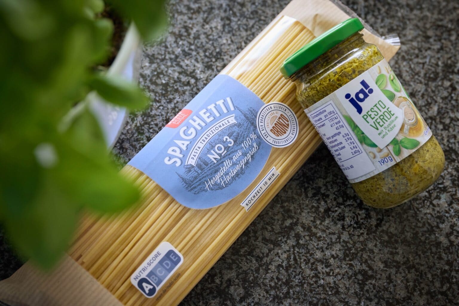 IDEE Living - Eine Packung Spaghetti liegt neben einem Glas mit grüner Pestosoße auf einer Steinfläche. In der Ecke ist der Teil einer grünen Pflanze zu sehen.