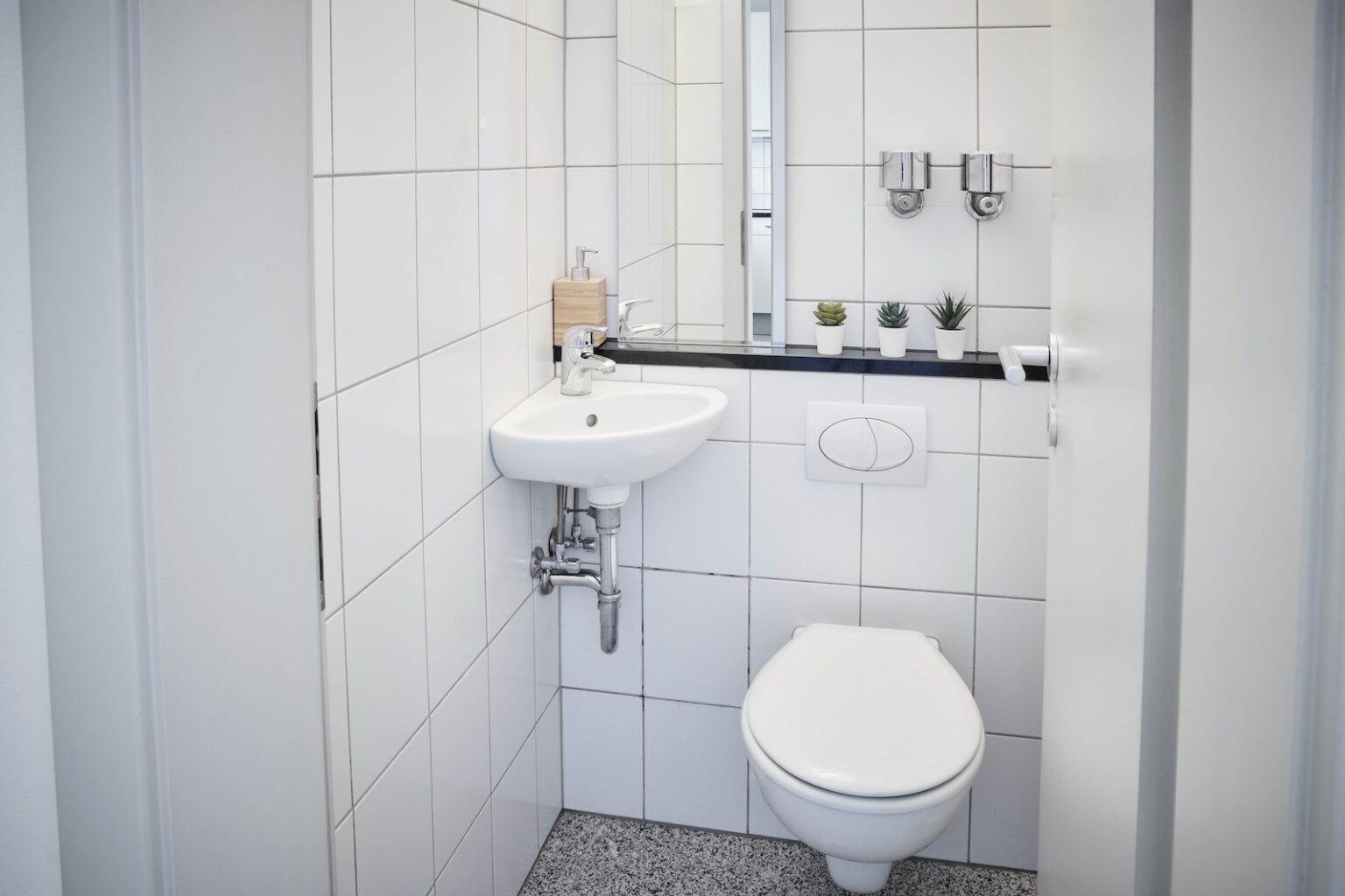 IDEE Living - Ein kleines, modernes Badezimmer mit weiß gefliesten Wänden verfügt über ein wandmontiertes Waschbecken und eine Toilette, einen rechteckigen Spiegel, einen Seifenspender und eine Reihe von drei kleinen Topfpflanzen auf einem schwarzen Regal.