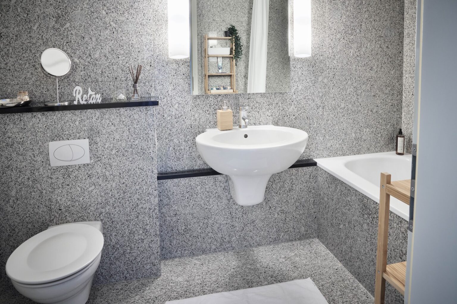 IDEE Living - Ein modernes Badezimmer mit einem wandmontierten Waschbecken, einer Toilette, einer Badewanne, einem kleinen Regal mit Dekorationsgegenständen, einem rechteckigen Spiegel und einem Handtuchhalter an einer grau gefliesten Wand.