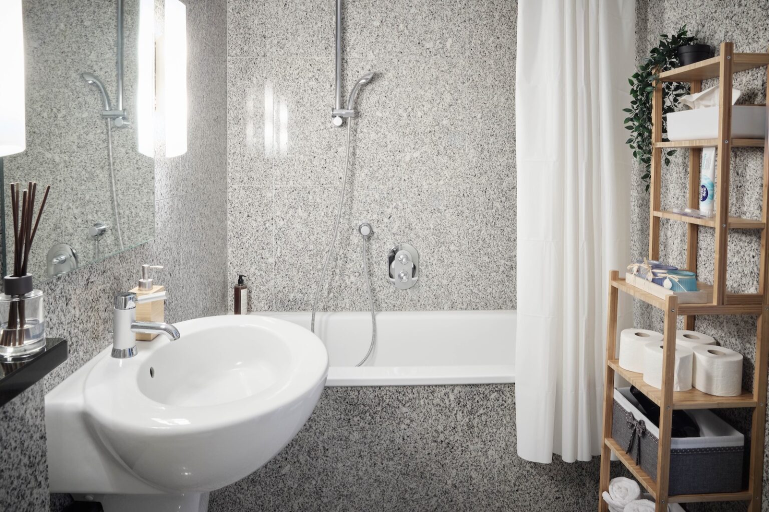 IDEE Living - Modernes Badezimmer mit Badewanne und Dusche, weißem Waschbecken, Wandspiegel und einem Holzregal mit Toilettenartikeln, Handtüchern und Toilettenpapier.