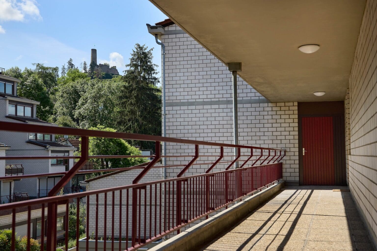 IDEE Living - Ein Balkon mit Ziegelwänden, rotem Geländer und Tür bietet Aussicht auf ein Gebäude und Grünflächen. In der Ferne ist vor einem blauen Himmel ein Turm zu erkennen.