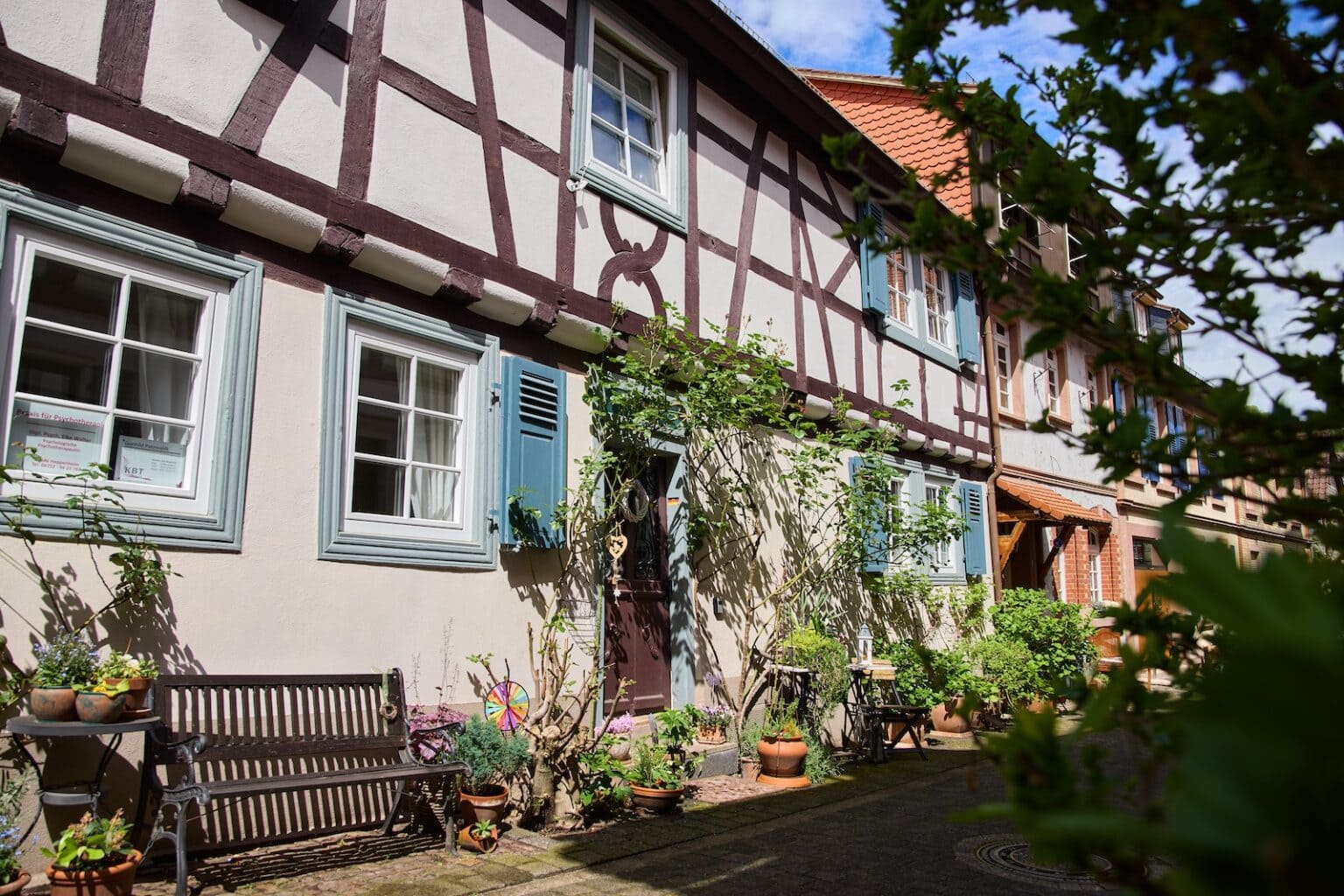 IDEE Living - Ein uriges Fachwerkhaus mit blauen Fensterläden und verschiedenen Topfpflanzen an der Fassade unter blauem Himmel.