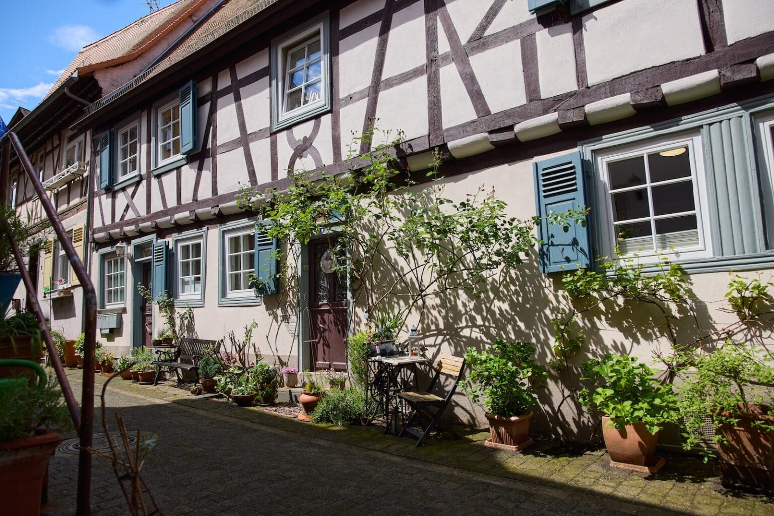 IDEE Living - Ein malerisches Fachwerkhaus mit blauen Fensterläden und Kletterpflanzen an der Fassade; ein kleiner Terrassenbereich bietet Topfpflanzen und einen Tisch mit Stühlen.