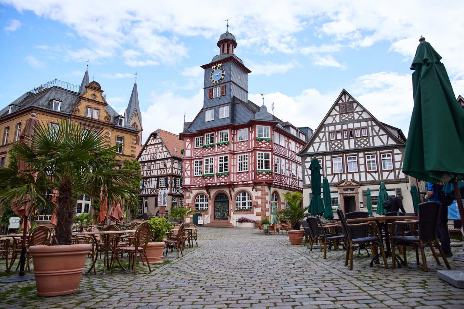 IDEE Living - Eine malerische Aussicht auf einen malerischen europäischen Stadtplatz mit Fachwerkhäusern, Sitzgelegenheiten im Freien bei Cafés, Topfpflanzen und einem Uhrturm unter einem teilweise bewölkten Himmel.