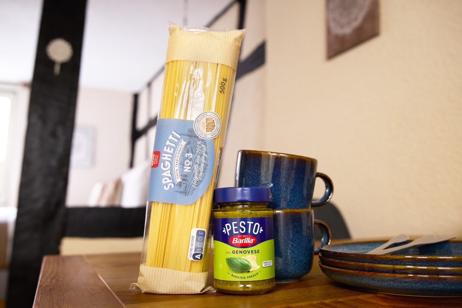 IDEE Living - Eine Packung Spaghetti und ein Glas Pestosoße auf einem Holztisch, im Hintergrund ein Set blauer Keramiktassen und -teller.