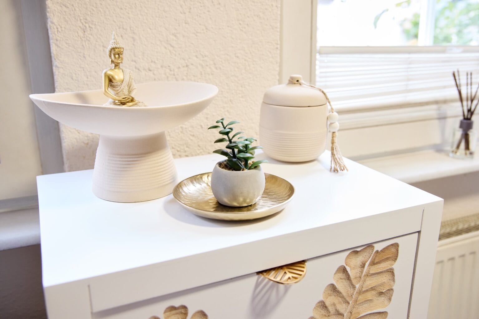 IDEE Living - Ein kleiner weißer Tisch mit einer dekorativen Schale, in der eine Buddha-Statue sitzt, einer kleinen Topfpflanze auf einem goldenen Teller, einem Behälter mit Deckel und Quaste und im Hintergrund einem Fenster mit Jalousien.