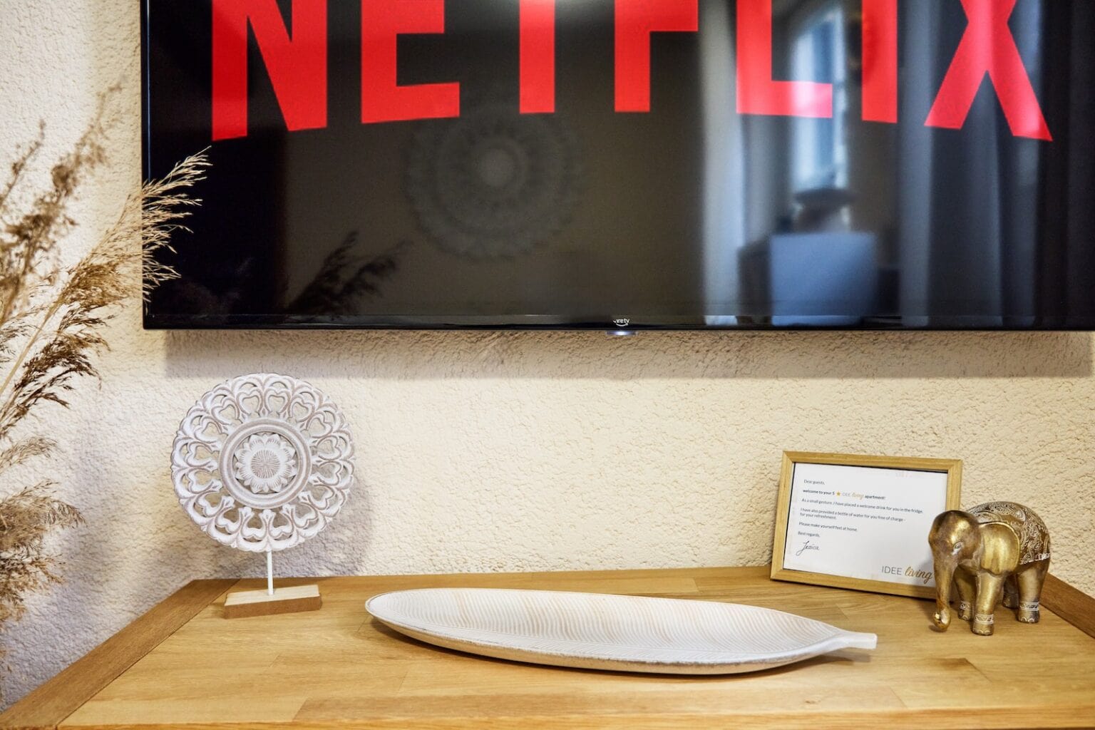 IDEE Living - Ein an der Wand montierter Fernseher mit dem Netflix-Logo über einer Holzkommode mit Dekorationsgegenständen, darunter ein weißer Zierteller, eine Keramikschale in Blattform, eine goldene Elefantenfigur und eine gerahmte Urkunde.