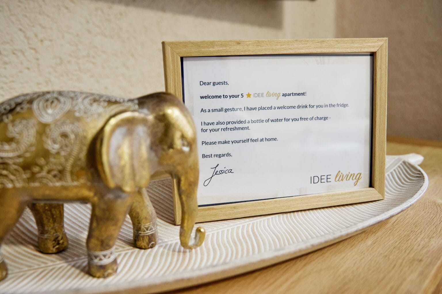 IDEE Living - Gerahmte Willkommenskarte neben einem goldenen dekorativen Elefanten auf einem Tablett. Die Karte enthält einen Gruß, erwähnt kostenloses Wasser und Getränke im Kühlschrank und ist mit „Jascha“ von IDEE Living signiert.