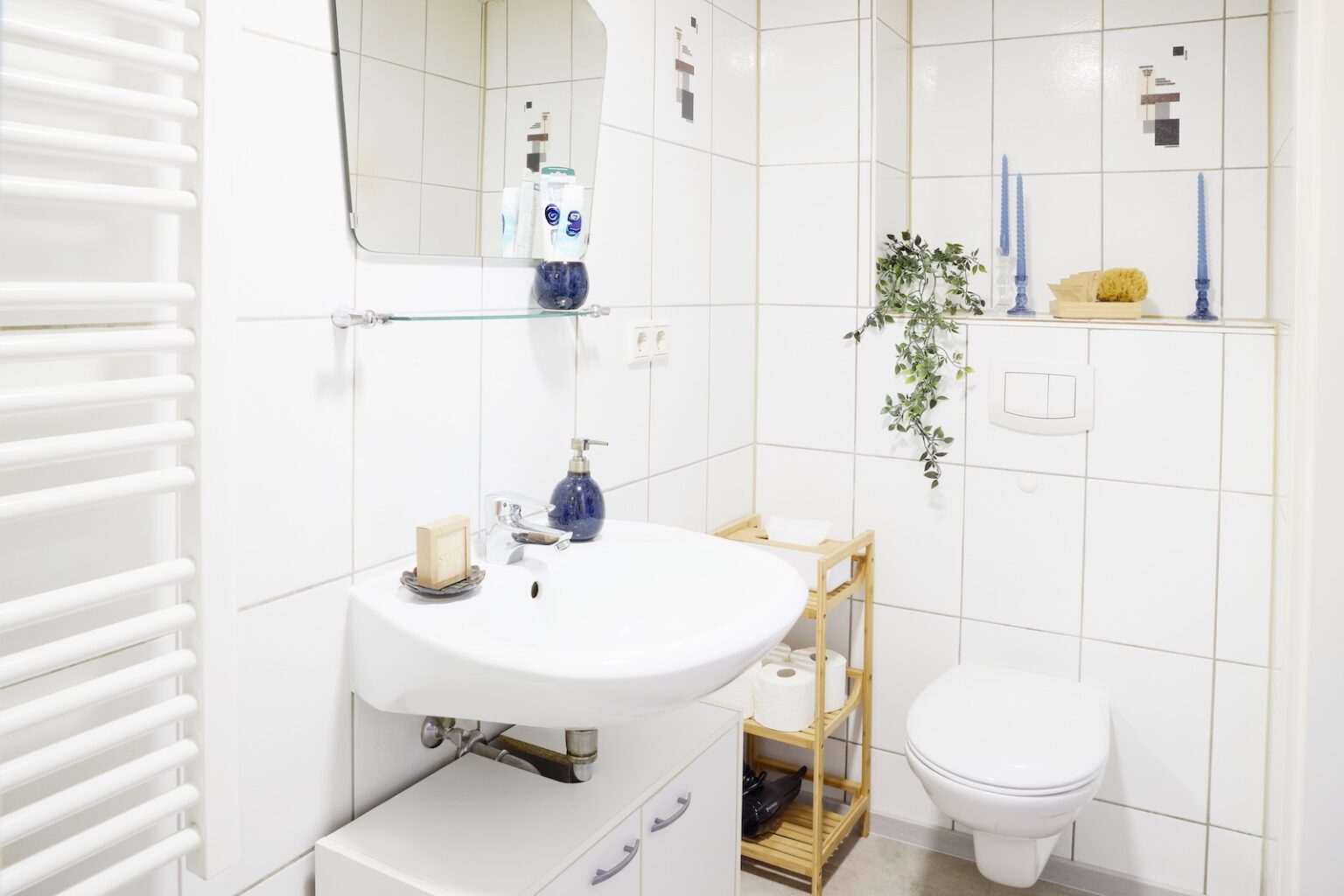 IDEE Living - Ein kleines Badezimmer mit weißem Waschbecken, Spiegel und Toilette. Es verfügt über ein Glasregal mit Toilettenartikeln, ein Holzregal mit Dekorationsgegenständen und blaue Kerzen auf einem Sims. Die Wände sind mit weißen Fliesen bedeckt.