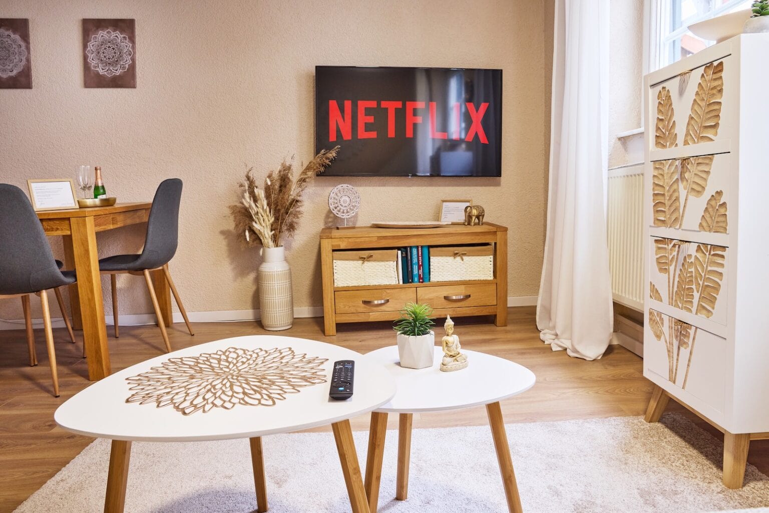 IDEE Living - Ein Wohnzimmer mit einem an der Wand montierten Fernseher mit dem Netflix-Logo, Holzmöbeln, einem weißen Couchtisch mit Fernbedienung und einer dekorativen Kommode.