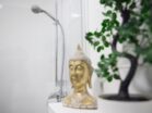 IDEE Living - Eine goldene Buddha-Statue steht neben einer Topfpflanze in einem Badezimmer mit Duschkopf und weißen Fliesen im Hintergrund.