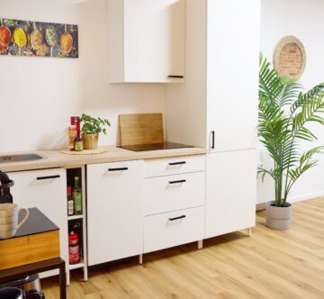 IDEE Living - Moderne Küche mit weißen Schränken, einer Spüle und einem Herd. Auf der Arbeitsplatte steht eine Topfpflanze. Es gibt einen kleinen Tisch mit einer Kaffeemaschine. Kunstwerke und ein Spiegel schmücken die Wände.