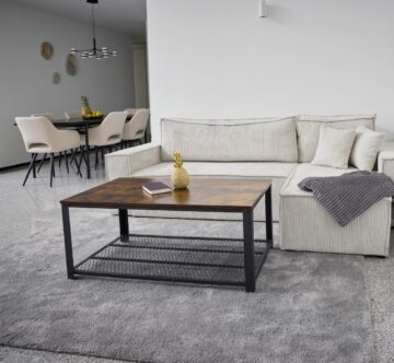 IDEE Living - Ein modernes Wohnzimmer mit einem hellgrauen Sofa, einem hölzernen Couchtisch und einem grauen Teppich. Im Hintergrund ist ein Essbereich mit beigen Stühlen zu sehen. Über dem Sofa liegt eine graue Decke.