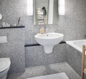 IDEE Living - Ein modernes Badezimmer mit einem wandmontierten Waschbecken, einer Toilette, einer Badewanne, einem kleinen Regal mit Dekorationsgegenständen, einem rechteckigen Spiegel und einem Handtuchhalter an einer grau gefliesten Wand.