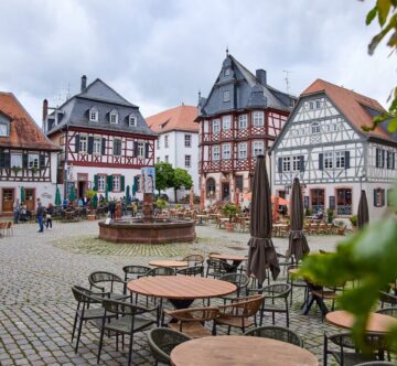 IDEE Living - Ein kopfsteingepflasterter Stadtplatz mit Sitzgelegenheiten im Freien, umgeben von Fachwerkhäusern mit roten und schwarzen Details.