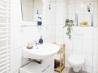 IDEE Living - Ein kleines Badezimmer mit weißem Waschbecken, Spiegel und Toilette. Es verfügt über ein Glasregal mit Toilettenartikeln, ein Holzregal mit Dekorationsgegenständen und blaue Kerzen auf einem Sims. Die Wände sind mit weißen Fliesen bedeckt.